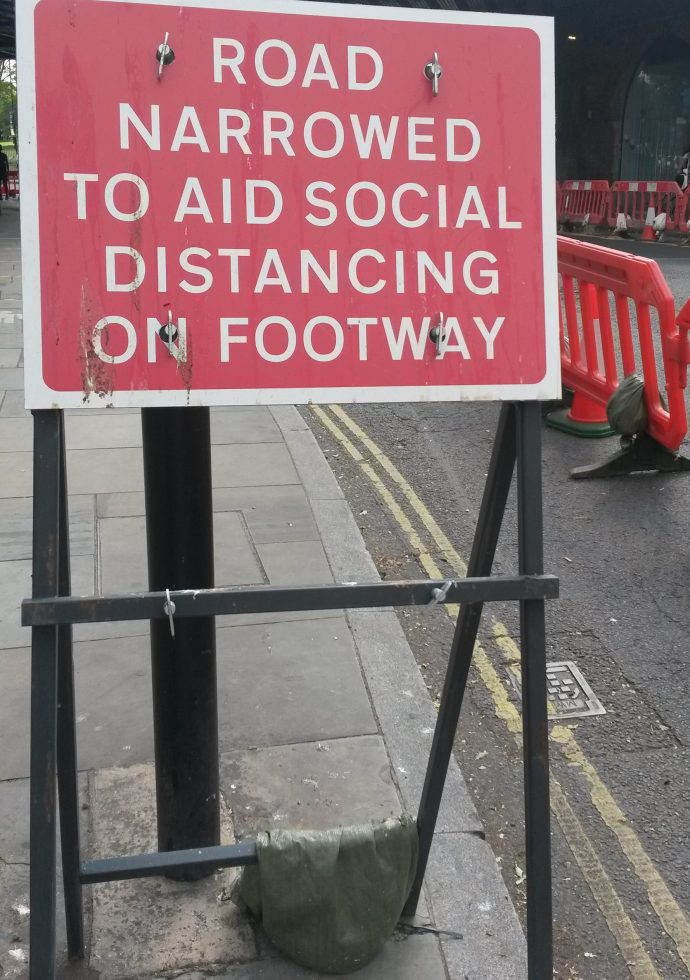 Sign on footpath saying road narrowed to aid social distancing on footway